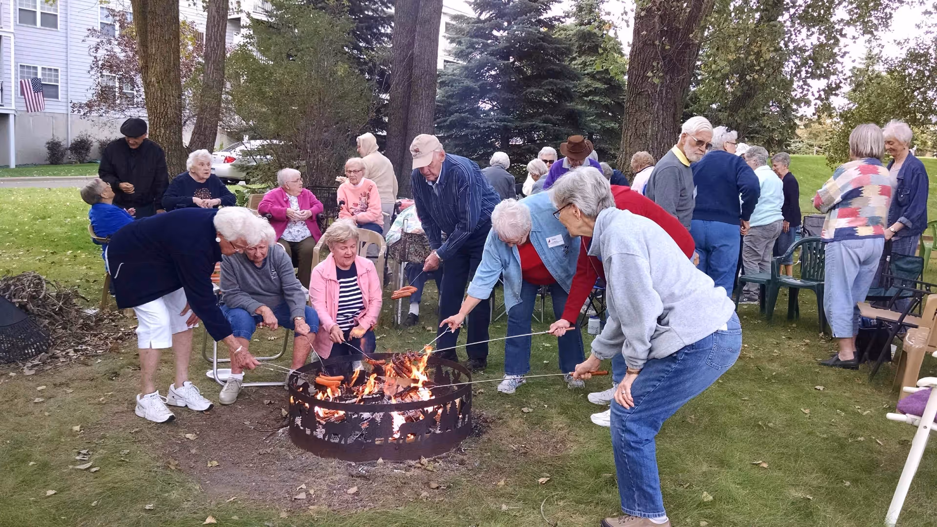 A group of elderly people gathered outdoors around a fire pit, roasting hot dogs on sticks. Some are seated while others stand, enjoying a social event on a grassy area with trees and a building in the background.