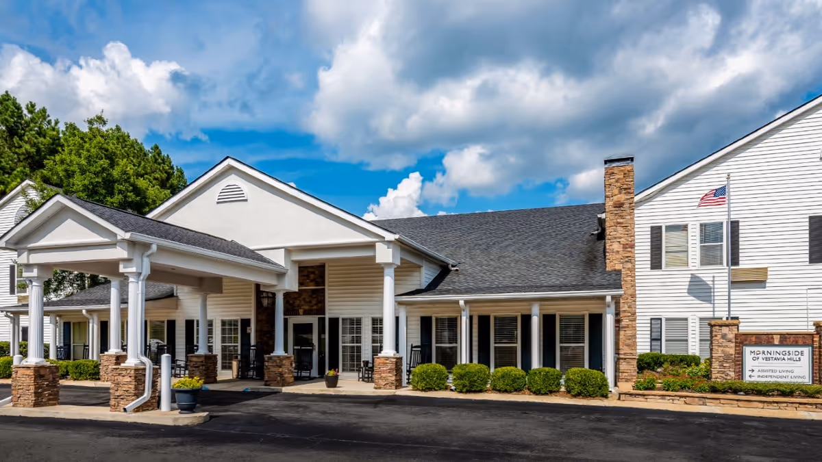 Exterior view of Morningside of Vestavia Hills senior living facility showing a white building with a covered entrance supported by white columns with stone bases, a driveway, green bushes, and an American flag on a flagpole under a partly cloudy blue sky.