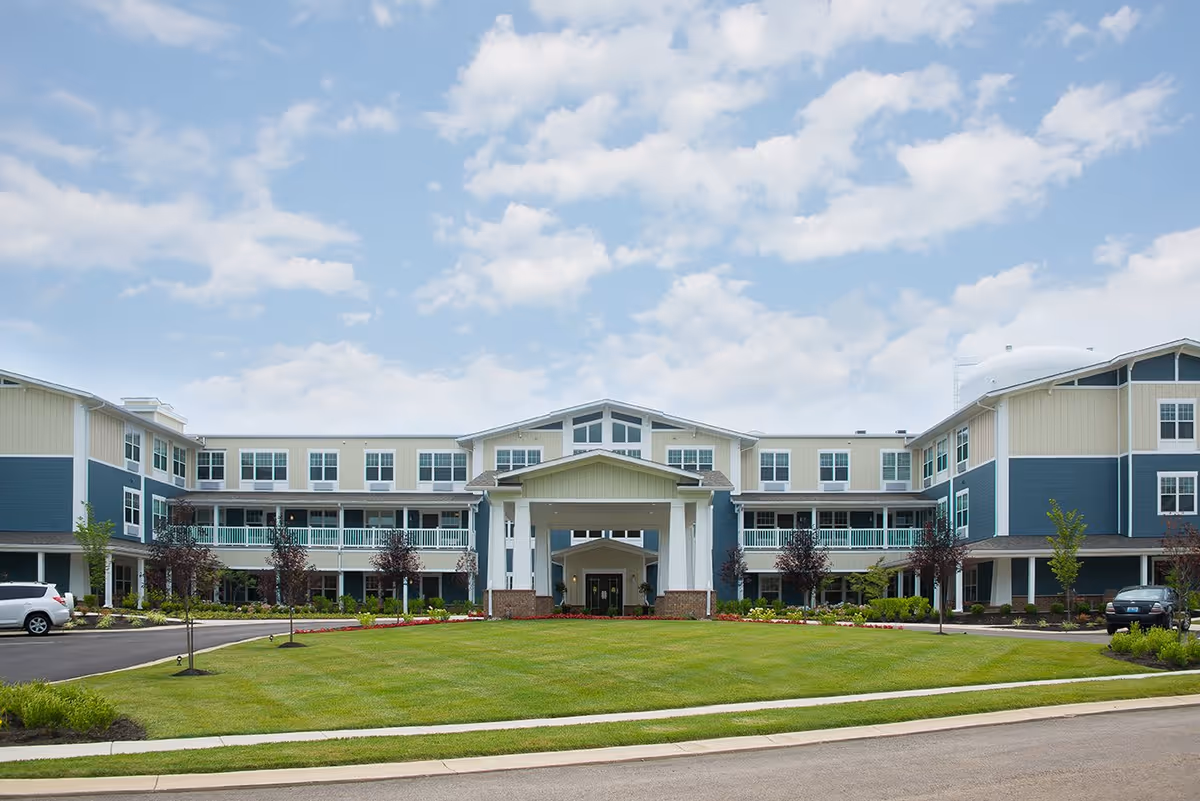 Front exterior view of a three-story senior living facility building named Traditions at Beaumont, featuring a large covered entrance, multiple windows, landscaped lawn, and a partly cloudy sky.