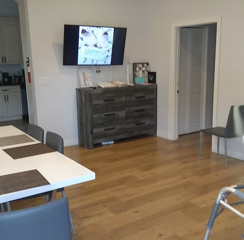 Interior view of a senior living facility room with a white table and gray chairs, a wooden dresser with items on top, a wall-mounted TV displaying an image, and a doorway leading to another room. The floor is wooden, and a walker is partially visible in the foreground.