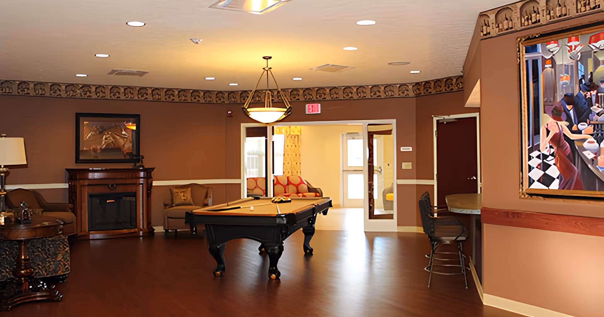 A cozy common area with a pool table in the center, surrounded by comfortable chairs and a small fireplace. The room has warm brown walls with decorative trim near the ceiling, a hanging light fixture above the pool table, and a colorful painting on the right wall. There is a bar area with stools on the right side and an exit door in the background leading to a brightly lit room with a sofa.