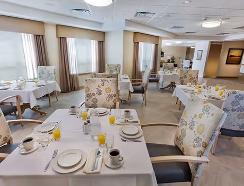 A bright and spacious dining room with multiple tables set for breakfast, featuring white tablecloths, plates, cups of coffee, glasses of orange juice, and floral upholstered chairs. Large windows with beige curtains allow natural light to fill the room.