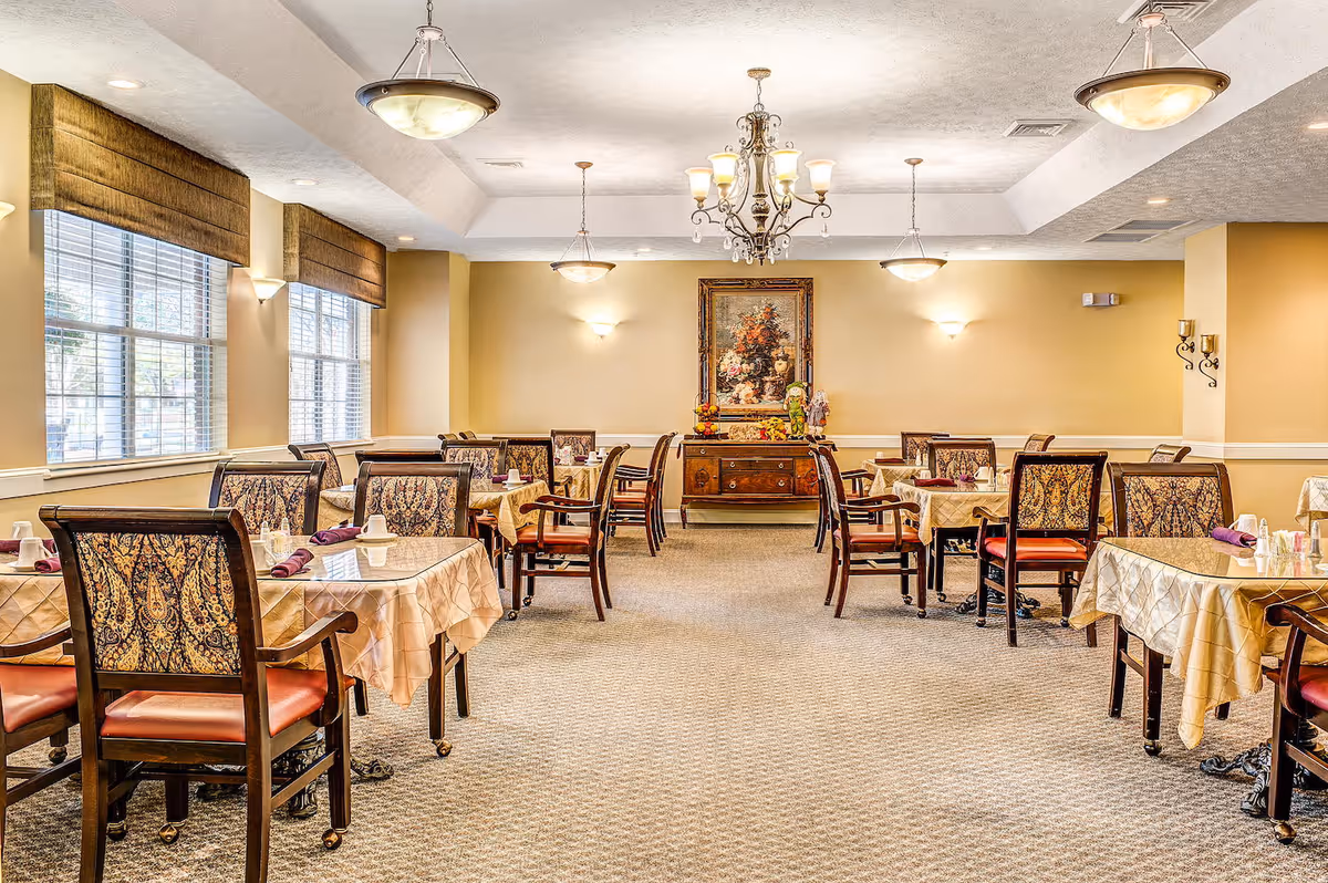 A well-lit dining room with multiple tables covered in beige tablecloths and set with folded purple napkins and cups. The chairs have patterned upholstery and wooden frames. Large windows with brown valances let in natural light. A chandelier and ceiling lights illuminate the room. A sideboard with decorative items and a framed floral painting is visible on the far wall.