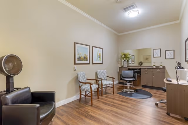 Small salon room with a styling chair and hooded dryer, two guest chairs, a mirror and cabinetry on wood floors.