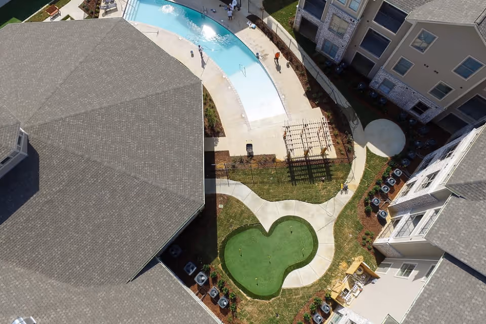 Aerial view of an outdoor area at The Claiborne at Hattiesburg Independent Living showing a kidney-shaped swimming pool, a small putting green, paved walkways, and surrounding buildings with gray roofs and beige walls.