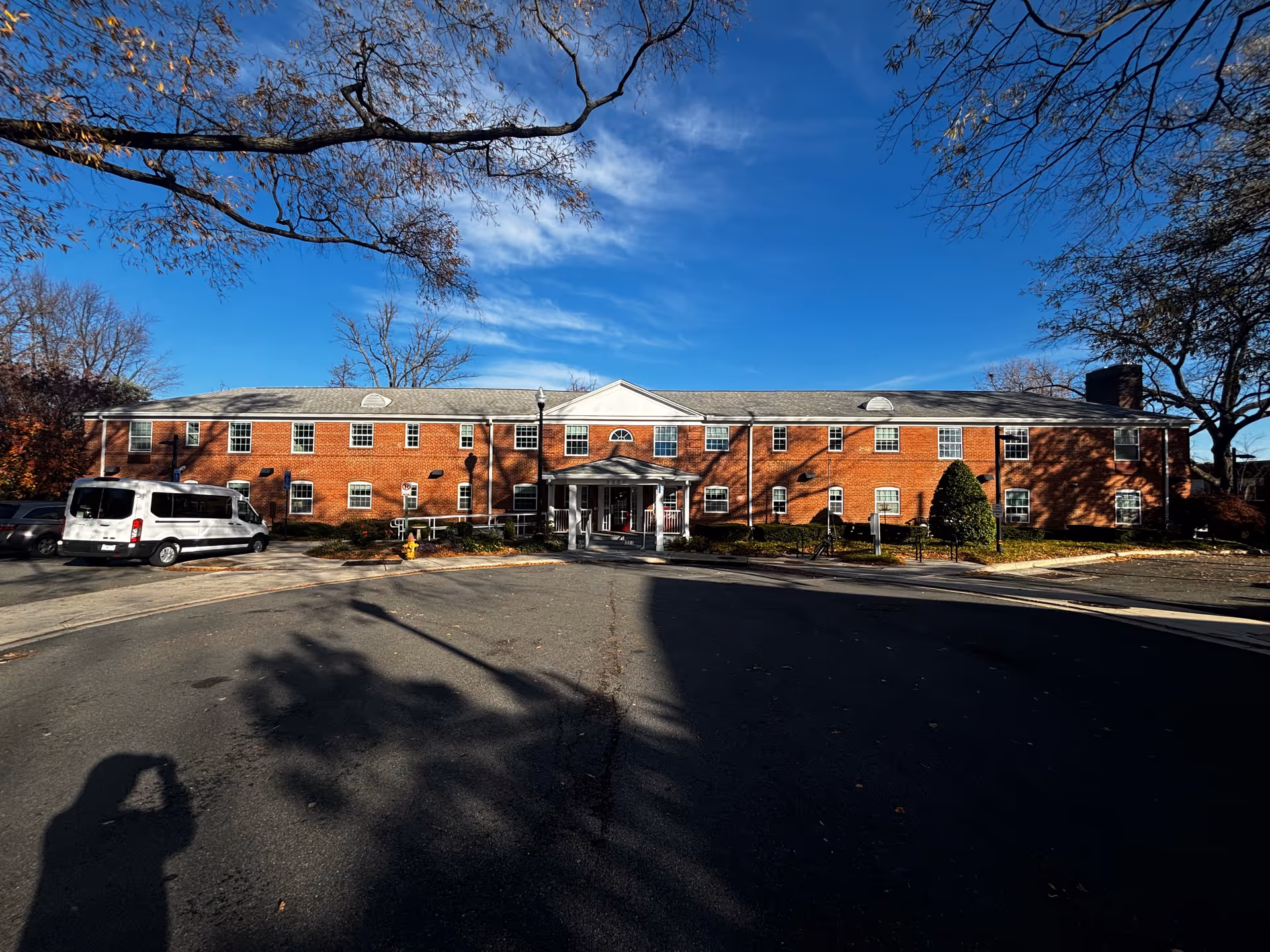 Front exterior of a two-story red brick assisted living building with a driveway, a white van parked to the left, and a bright blue sky overhead.