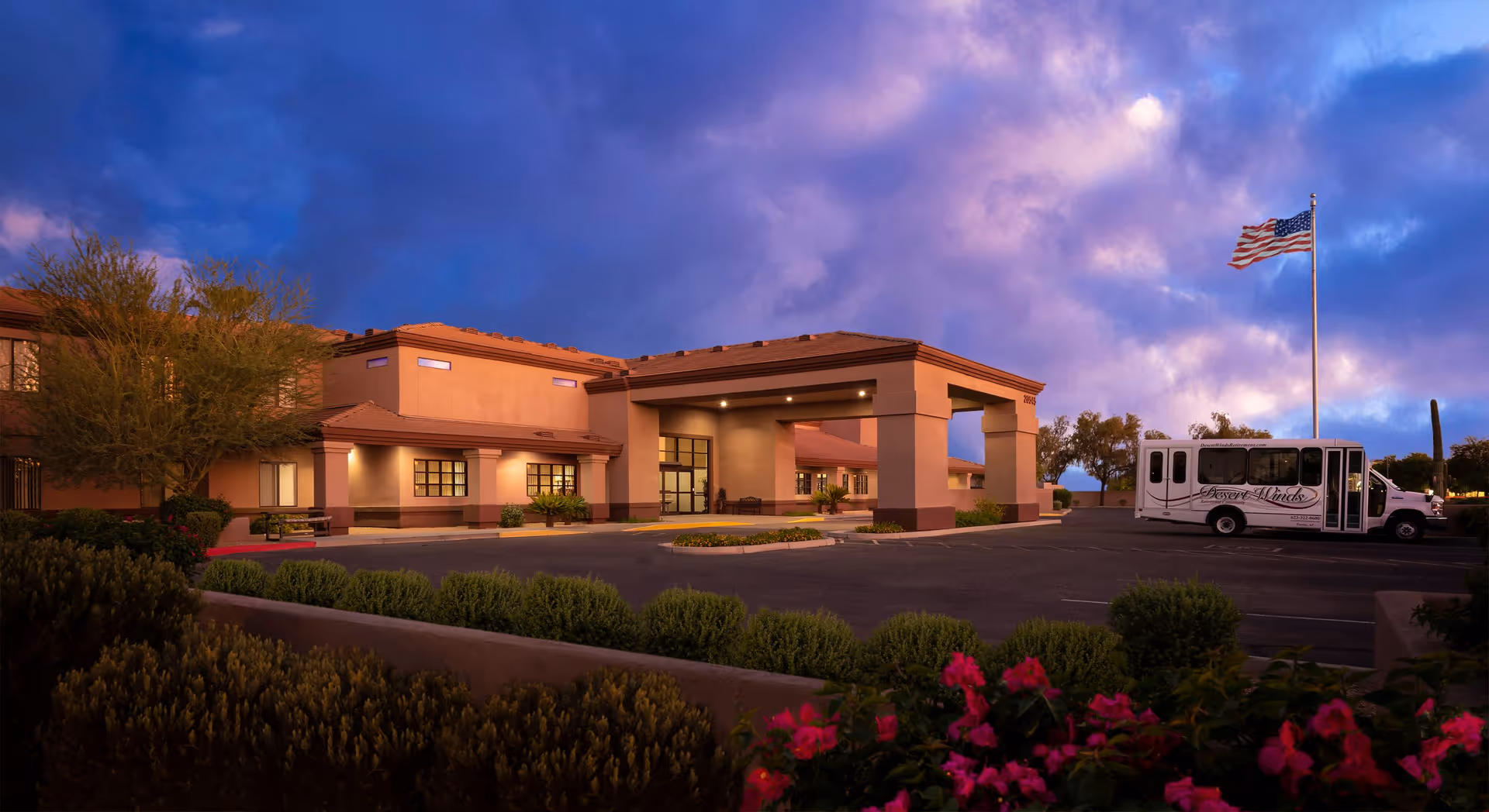 Exterior view of Desert Winds Assisted Living facility at dusk with a large entrance canopy, landscaped bushes and flowers in the foreground, an American flag on a tall flagpole, and a white shuttle bus parked in the lot.