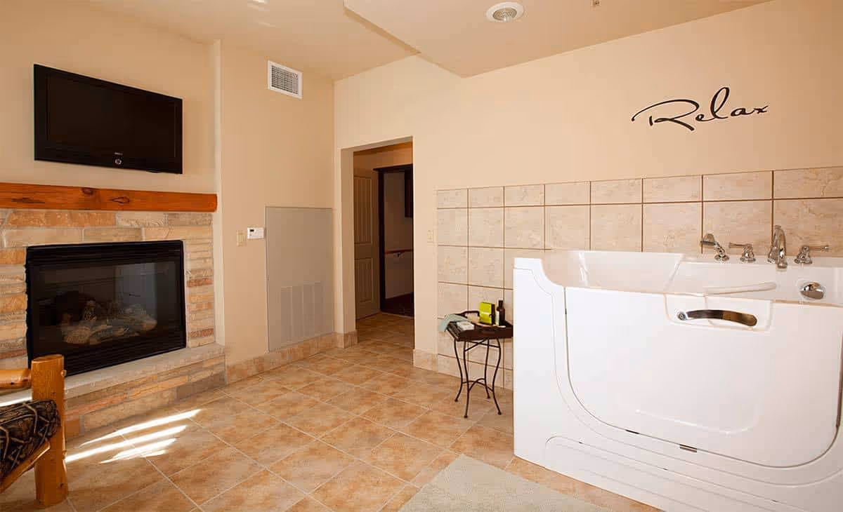 A tiled room featuring a white walk-in bathtub with faucets and a 'Relax' wall decal, a stone fireplace with a mounted TV, and a small side table.