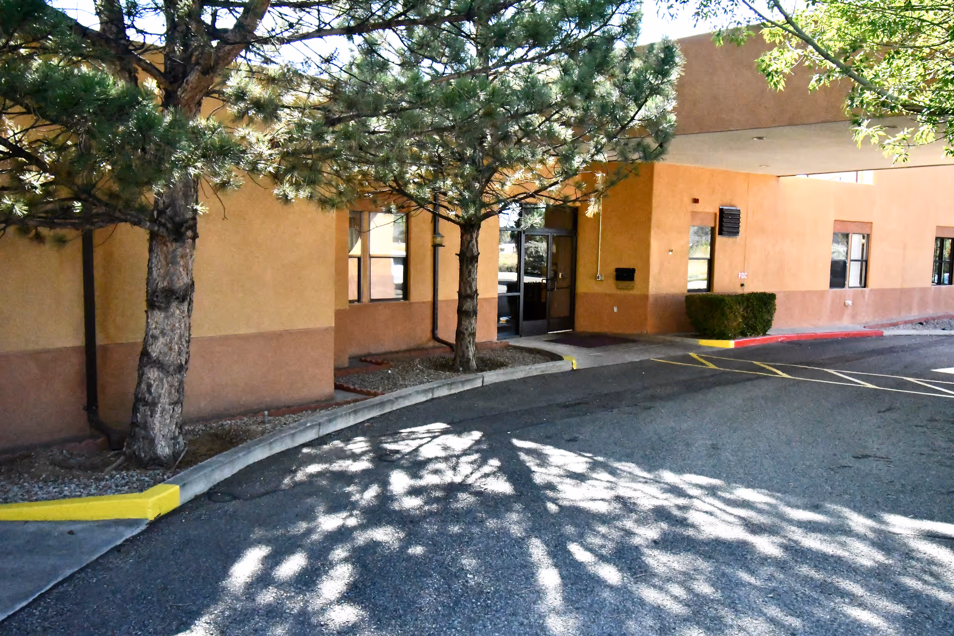 Exterior view of a building entrance with a covered drop-off area, surrounded by trees casting shadows on the pavement. The building has a beige and brown stucco finish with several windows and a glass door entrance.