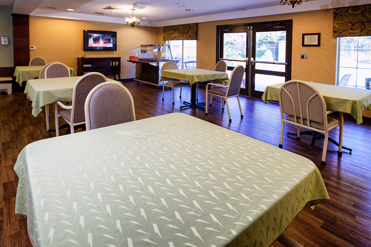 Interior view of a dining room in a senior living facility with several tables covered with light green tablecloths and chairs arranged around them. There is a television mounted on the wall, a serving station, large windows, and double glass doors letting in natural light.