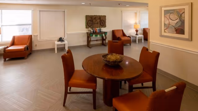 A cozy common area with a round wooden table surrounded by four brown chairs. In the background, there are two brown armchairs near windows with white blinds, a small white side table with a lamp, and a decorative console table with a bicycle wall art above it. The walls are light-colored with a framed abstract painting on the right.