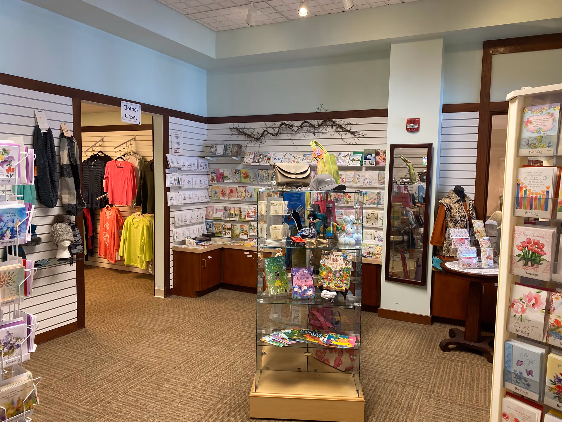 Interior view of a small gift shop area in a senior living facility with greeting cards displayed on racks and shelves, a glass display case with various items including books and stuffed animals, and a clothes closet with hanging sweaters and jackets.