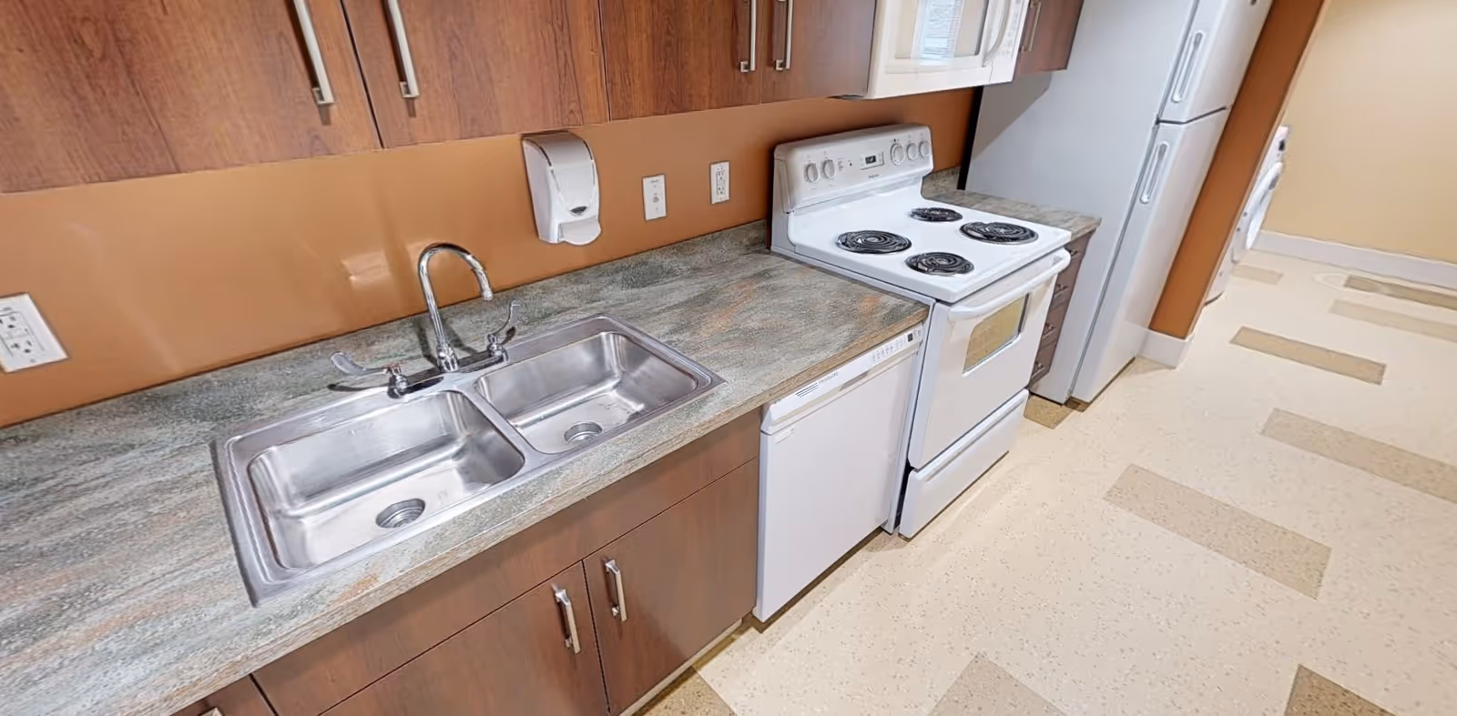 A kitchen area with a double stainless steel sink, a white electric stove with four coil burners, a white dishwasher, a white refrigerator, and wooden cabinets above and below the countertop. The floor has a beige and light brown patterned linoleum.
