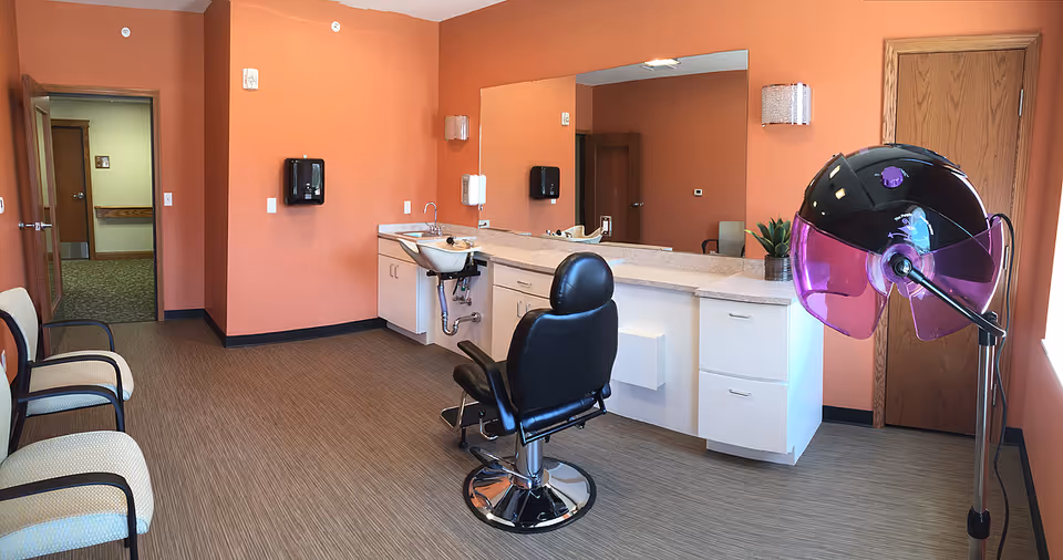 A salon room with peach-colored walls featuring a black salon chair in front of a counter with a sink and cabinets. There is a large mirror above the counter, a hair dryer with a purple hood on a stand, and three beige chairs with dark armrests along one wall. A wooden door and an open doorway leading to a hallway are visible.