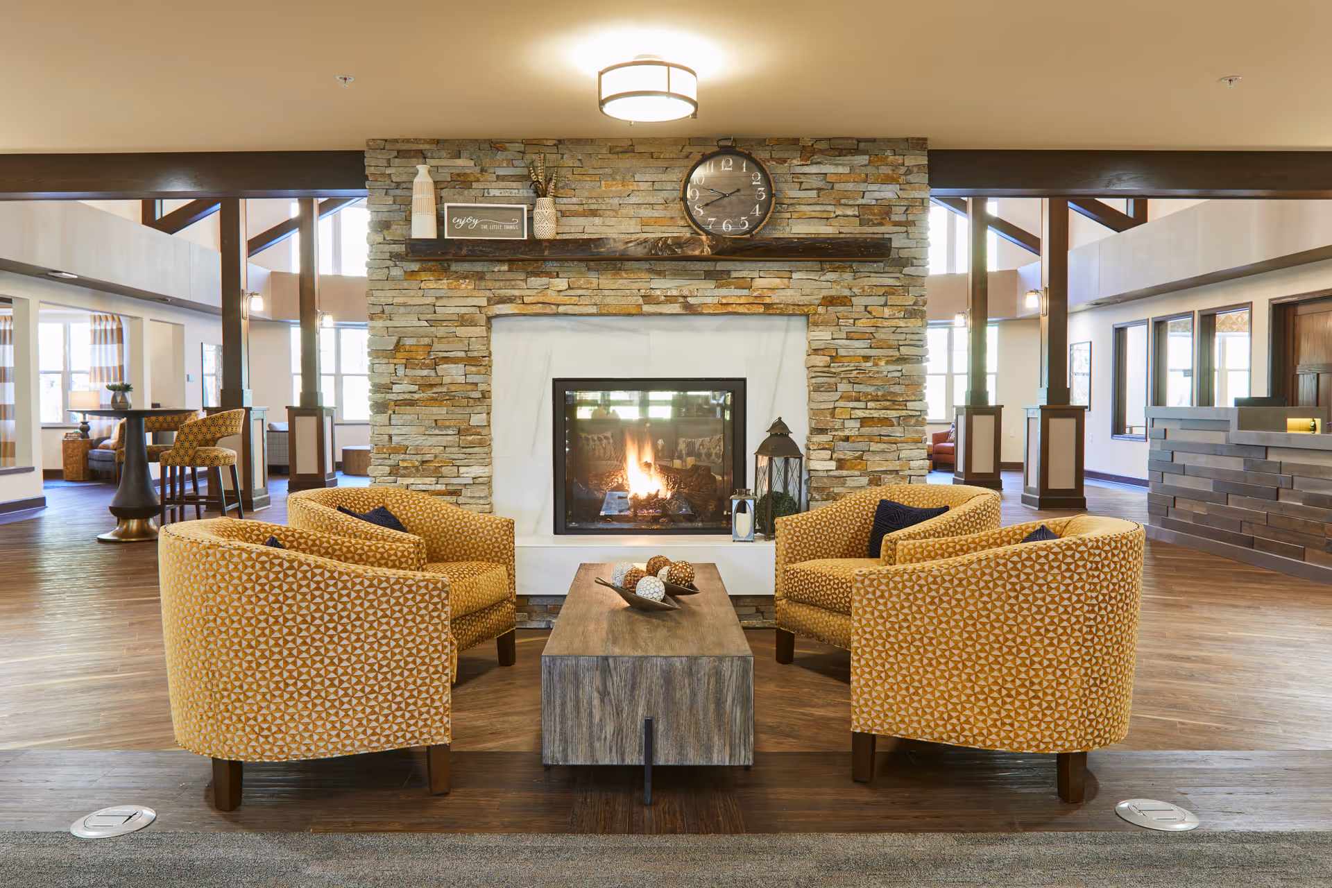 Cozy senior living lounge with a central stone fireplace, four yellow patterned armchairs arranged around a wooden coffee table, and a reception area in the background.