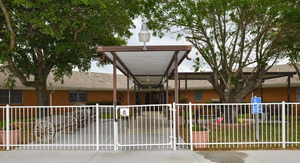 Entrance to a single-story brick building with a covered walkway supported by metal posts. The walkway is flanked by a white metal fence with a gate in the center. There are large trees on either side of the entrance and potted plants near the fence. A blue handicap parking sign is visible on the right side near the fence.