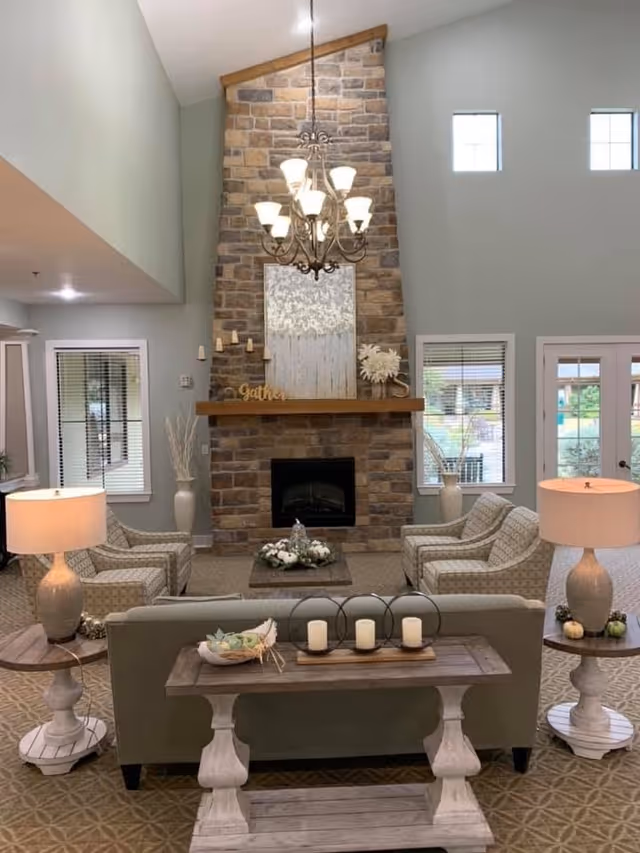 A cozy living room with a tall stone fireplace as the focal point. The room features a chandelier hanging from the high ceiling, two armchairs and a sofa arranged around a coffee table with decorative items. There are two table lamps on side tables flanking the sofa, windows with blinds, and a door leading outside. The decor includes candles, plants, and a wooden mantel with decorative letters spelling 'gather'.