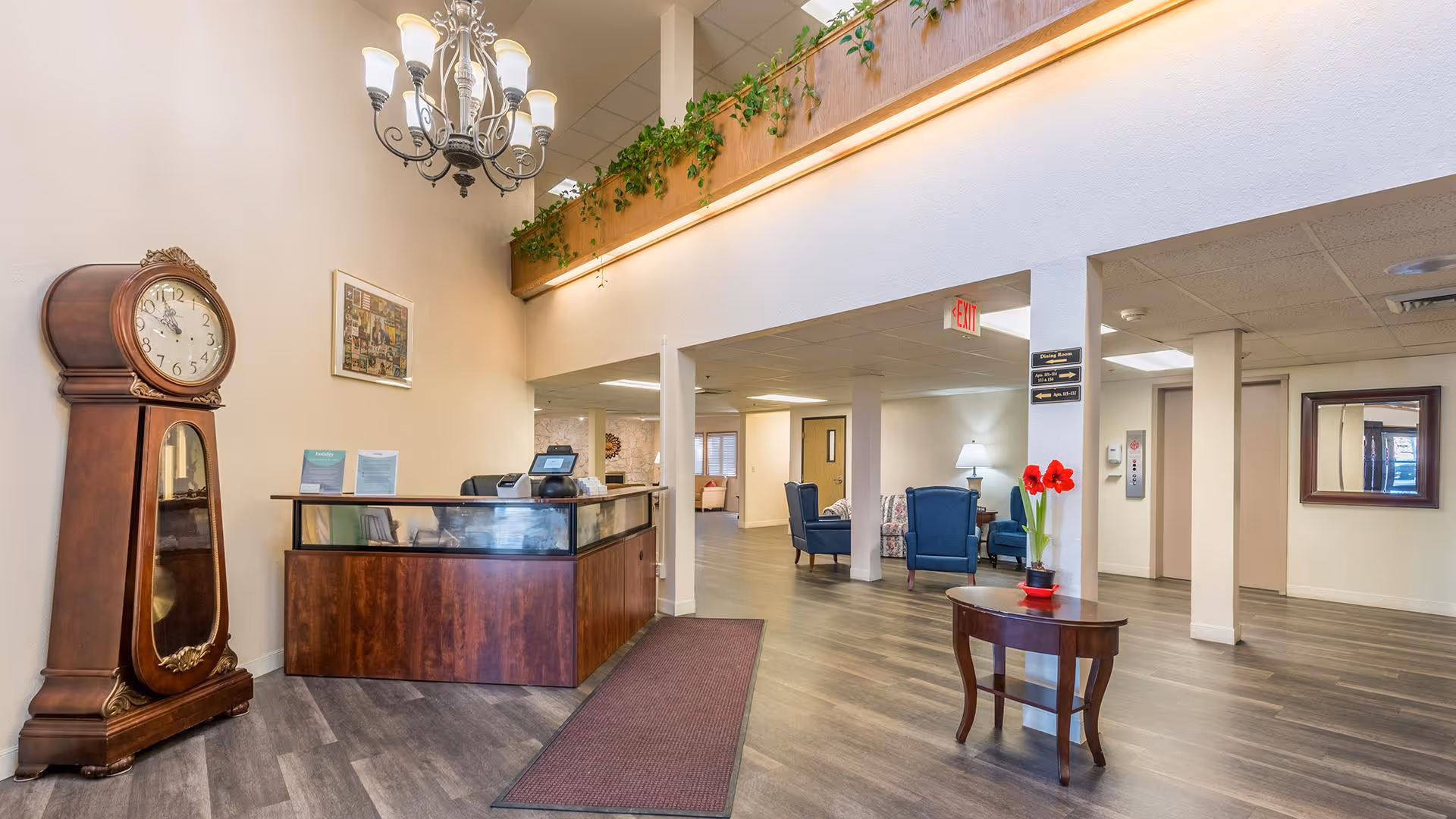 Interior view of a senior living facility lobby with a wooden reception desk, a large grandfather clock, a chandelier, and a small round table with a red flower pot. The space has wood flooring, white walls, and a high ceiling with plants along a ledge above the reception area. There are blue armchairs and a lamp visible in the background near an elevator and hallway.