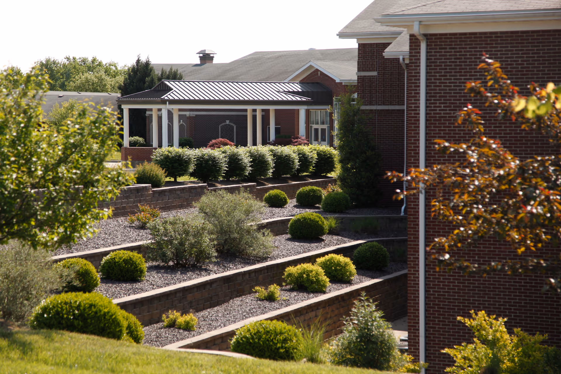 Terraced garden with neatly trimmed bushes and shrubs in front of a brick building with a covered walkway, surrounded by trees and greenery under a clear sky.