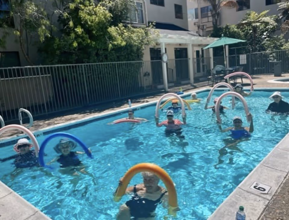 A group of elderly people in a swimming pool holding colorful pool noodles, participating in a water exercise class. The pool is outdoors, surrounded by a fence, trees, and a building in the background.