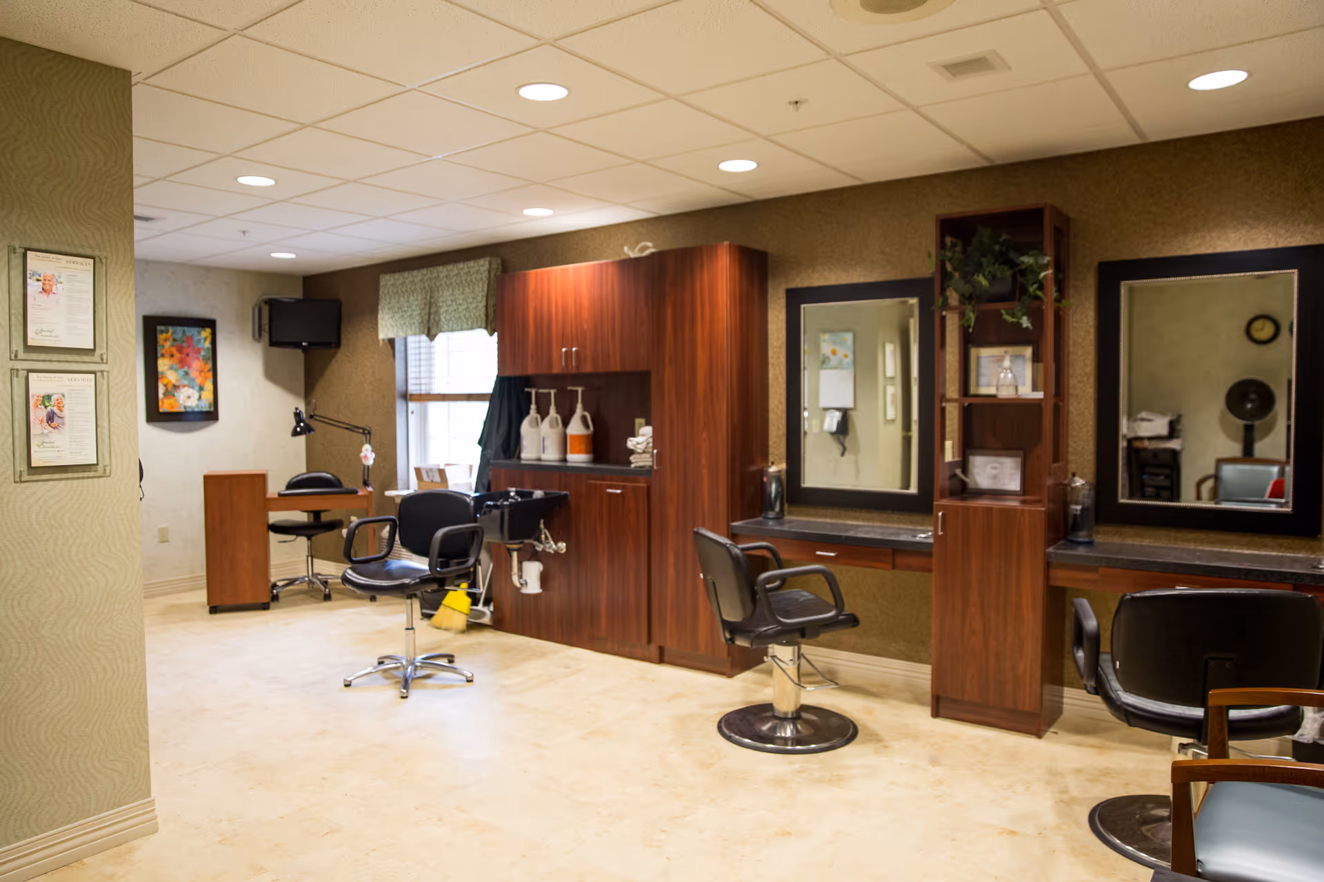 Interior salon area with styling chairs, mirrors, wooden cabinets, and a small desk in a senior living facility.