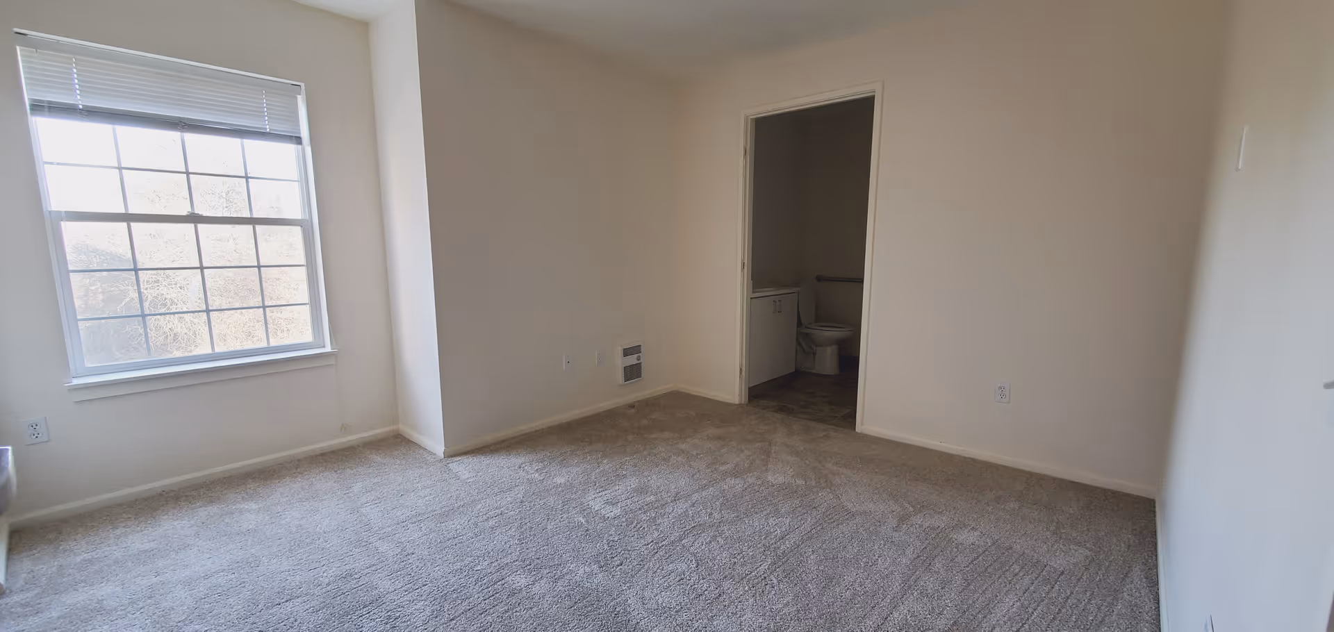 Empty room with beige carpet and white walls, a large window with blinds on the left side, and an open doorway leading to a bathroom with a visible toilet and sink.