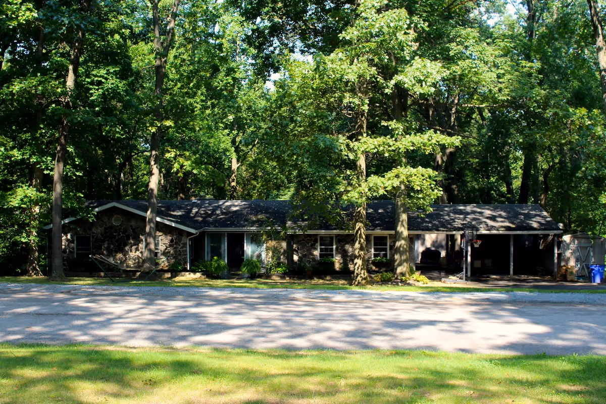 A single-story stone house surrounded by tall trees with a gravel driveway in front. The house has a covered carport on the right side and a hammock set up between two trees on the left side. The area is shaded by the dense foliage.