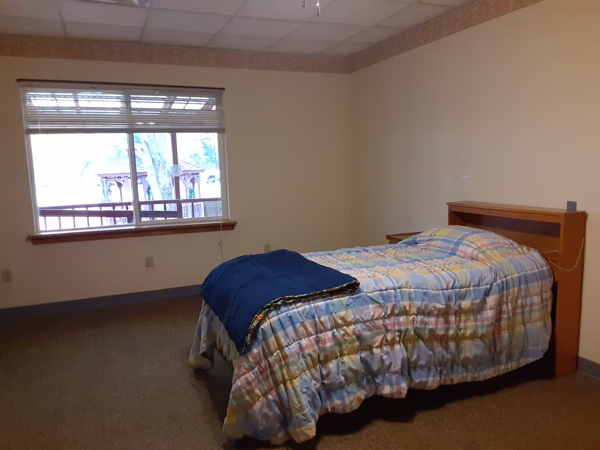 A simple bedroom with a single bed covered in a colorful patchwork quilt and a dark blue blanket folded at the foot. The bed has a wooden headboard with a small nightstand beside it. There is a large window with blinds partially open, showing an outdoor view with trees and a wooden structure.