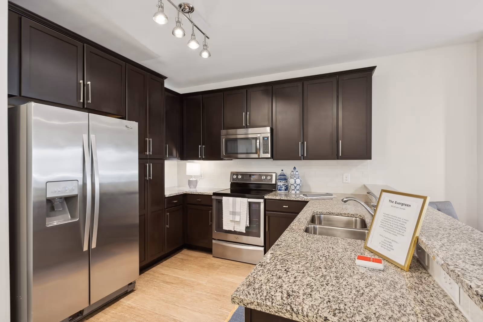 Modern kitchen with dark wood cabinets, stainless steel refrigerator, stove, and microwave. Granite countertops with a double sink and a framed sign on the counter. Light wood flooring and white walls.