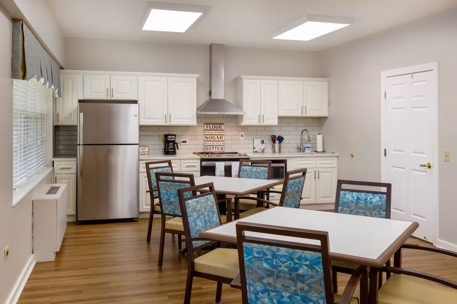 Communal dining area with several tables and patterned chairs facing a small kitchen with white cabinets and a stainless steel refrigerator.