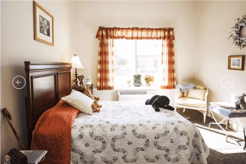 A cozy bedroom with a wooden headboard bed covered in a white quilt with a circular pattern and an orange blanket. There are two stuffed animals on the bed, a teddy bear and a black dog. A window with orange plaid curtains lets in natural light. To the right, there is a cushioned chair with a folded blanket on it and a small table with books and a decorative item. A walking cane leans against the wall near the bed, and a framed picture hangs above the headboard.