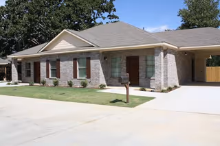 Single-story brick apartment building with a manicured lawn, driveway, mailbox, and a carport under a clear blue sky.
