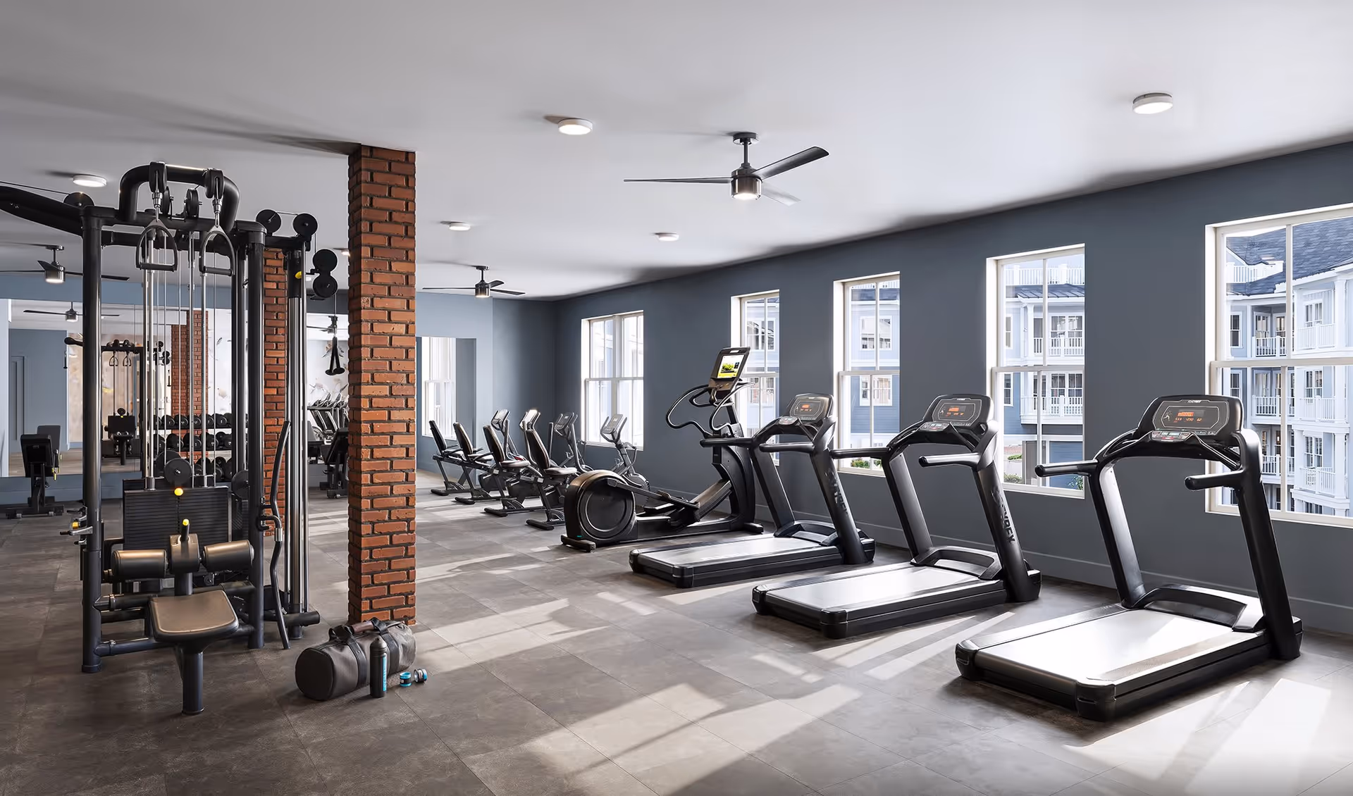Bright fitness room with treadmills, cardio machines, and weight equipment arranged near large windows.