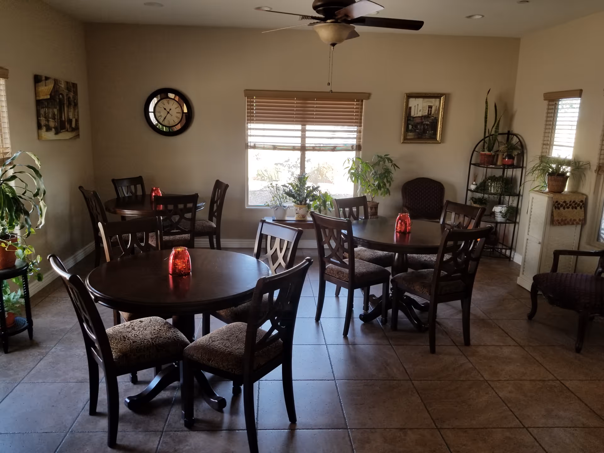 A cozy dining room with three round wooden tables, each surrounded by four cushioned chairs. Each table has a red candle holder in the center. The room has beige walls, tiled floor, and several windows with blinds partially open, letting in natural light. There are various potted plants on stands and shelves, a wall clock, and framed artwork on the walls. A ceiling fan with a light fixture is mounted on the ceiling.