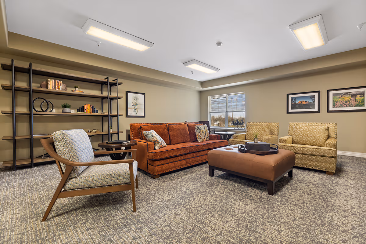 A cozy senior living facility lounge area with a rust-colored sofa, two patterned armchairs, a cushioned ottoman with a tray, a wooden side table, and a large open bookshelf against the wall. The room has beige walls, carpeted flooring, framed pictures on the walls, and a window letting in natural light.