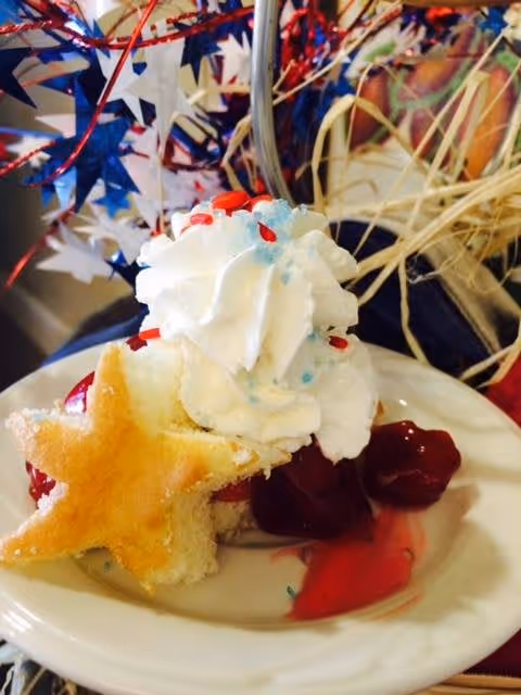 A dessert plate featuring a slice of cake topped with whipped cream and red and blue sprinkles, garnished with a star-shaped cookie and a serving of cherry topping. In the background, there are red, white, and blue decorative elements.