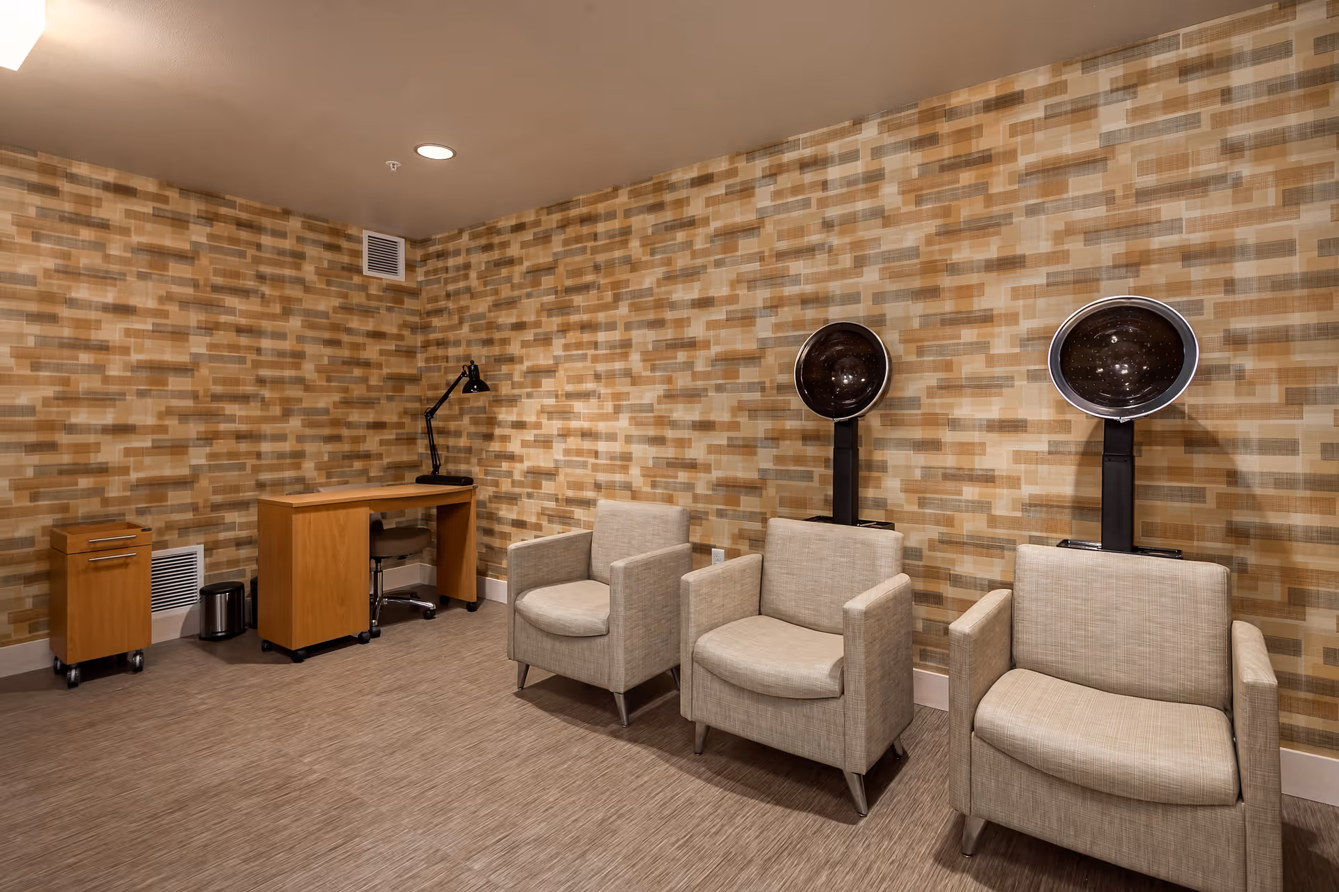 Interior room with three beige armchairs, each positioned under a black hair dryer hood. To the left, there is a wooden desk with a black adjustable lamp and a rolling cabinet. The walls have a patterned wallpaper in shades of brown and beige, and the floor is covered with a light brown carpet.