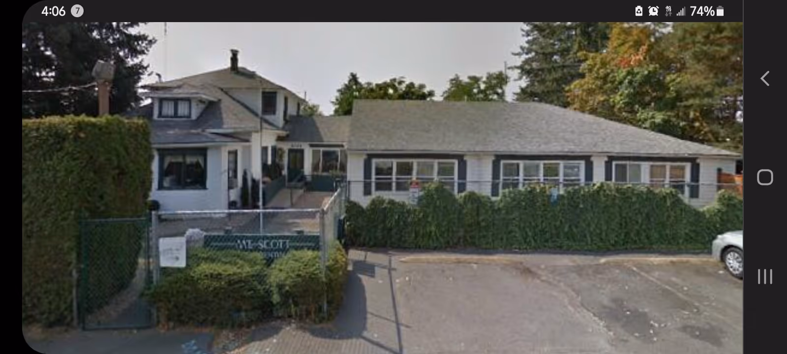 Front exterior of a single-story residential care building with a gated entrance, tall hedges and a small parking lot.