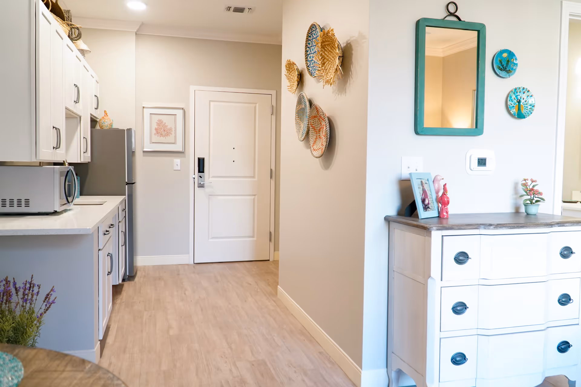 Interior view of a senior living facility kitchen area with white cabinets, a microwave, and a refrigerator. The floor is light wood, and there is a white door at the end of the hallway. On the right wall, decorative woven baskets and a teal-framed mirror hang above a white dresser with black handles. Small decorative items and a potted plant are on top of the dresser.