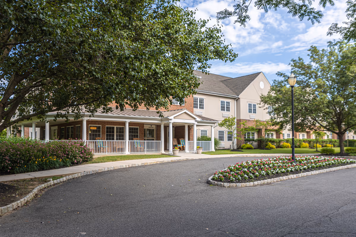 Front exterior of a senior living building with a covered porch, circular driveway, trees, and landscaped flower beds.