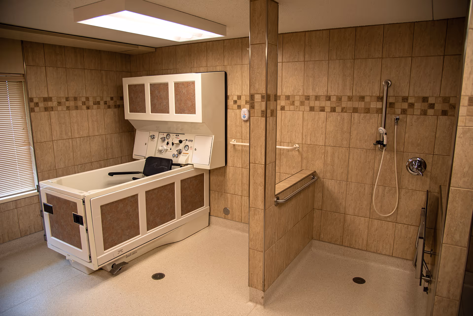 Accessible tiled bathroom with a medical bathing tub, wall-mounted controls, and an open shower area with grab bars.