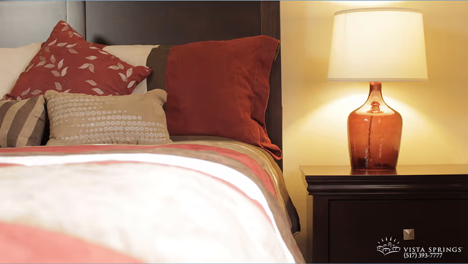 Close-up view of a bed with multiple decorative pillows in shades of red, beige, and brown, next to a dark wooden nightstand with a red glass lamp and a beige lampshade, softly illuminating the area.