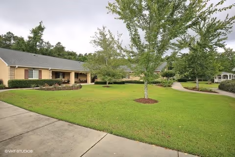 A well-maintained outdoor area of a senior living facility with green grass, several trees, a paved walkway, and a beige building with a gray roof in the background under a cloudy sky.