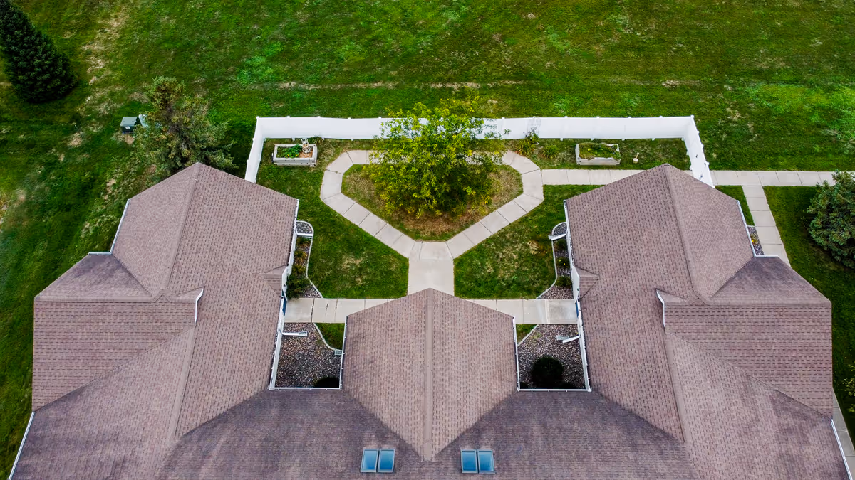 Aerial view of a senior living facility showing the roof of a building with multiple sections and a central courtyard with a tree and paved walkways forming a heart shape, surrounded by green grass.