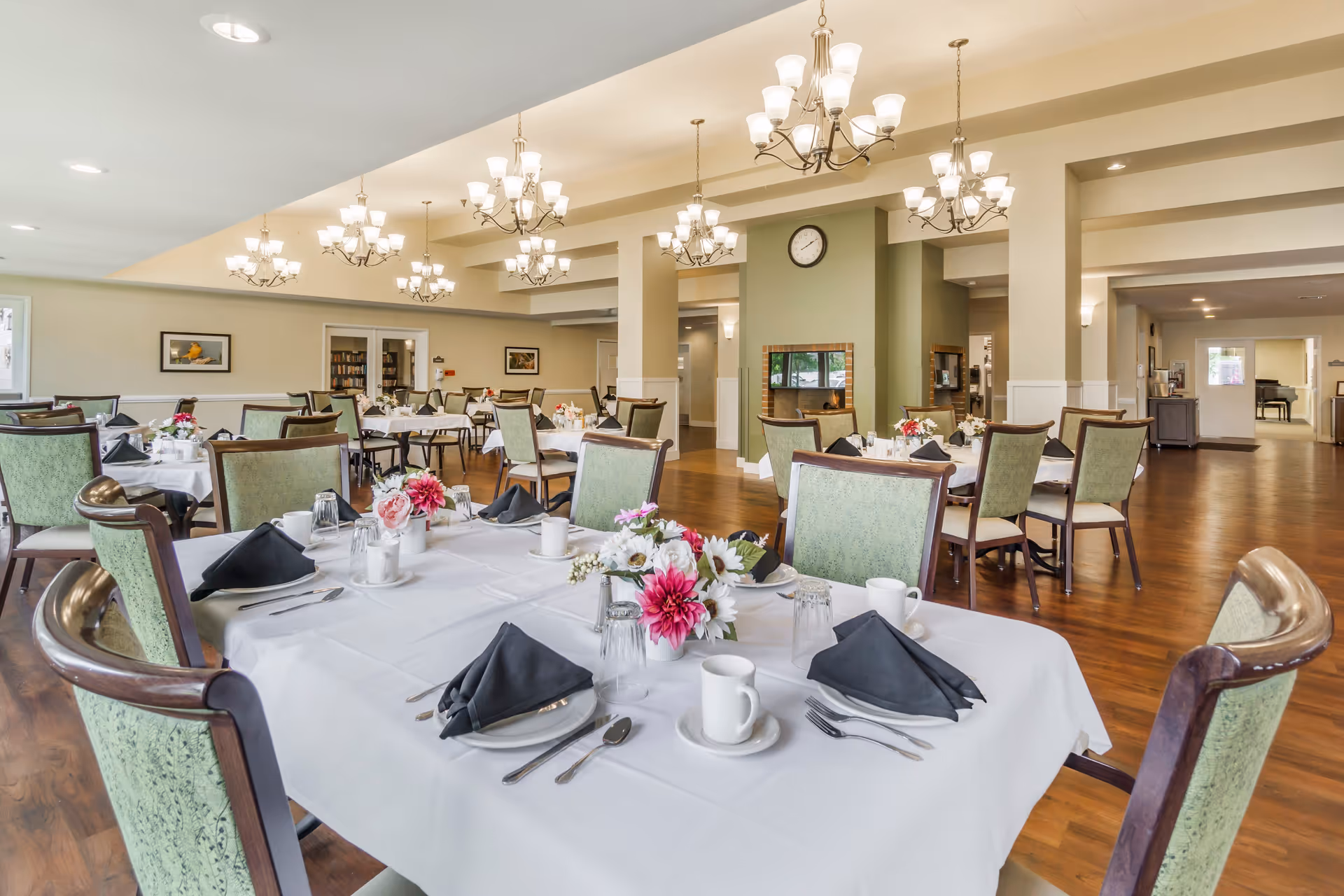 A spacious dining room with multiple tables covered in white tablecloths, each set with black napkins, cups, utensils, and floral centerpieces. The room features green upholstered chairs, wooden flooring, and several chandeliers hanging from the ceiling. A clock is mounted on a green accent wall in the background.