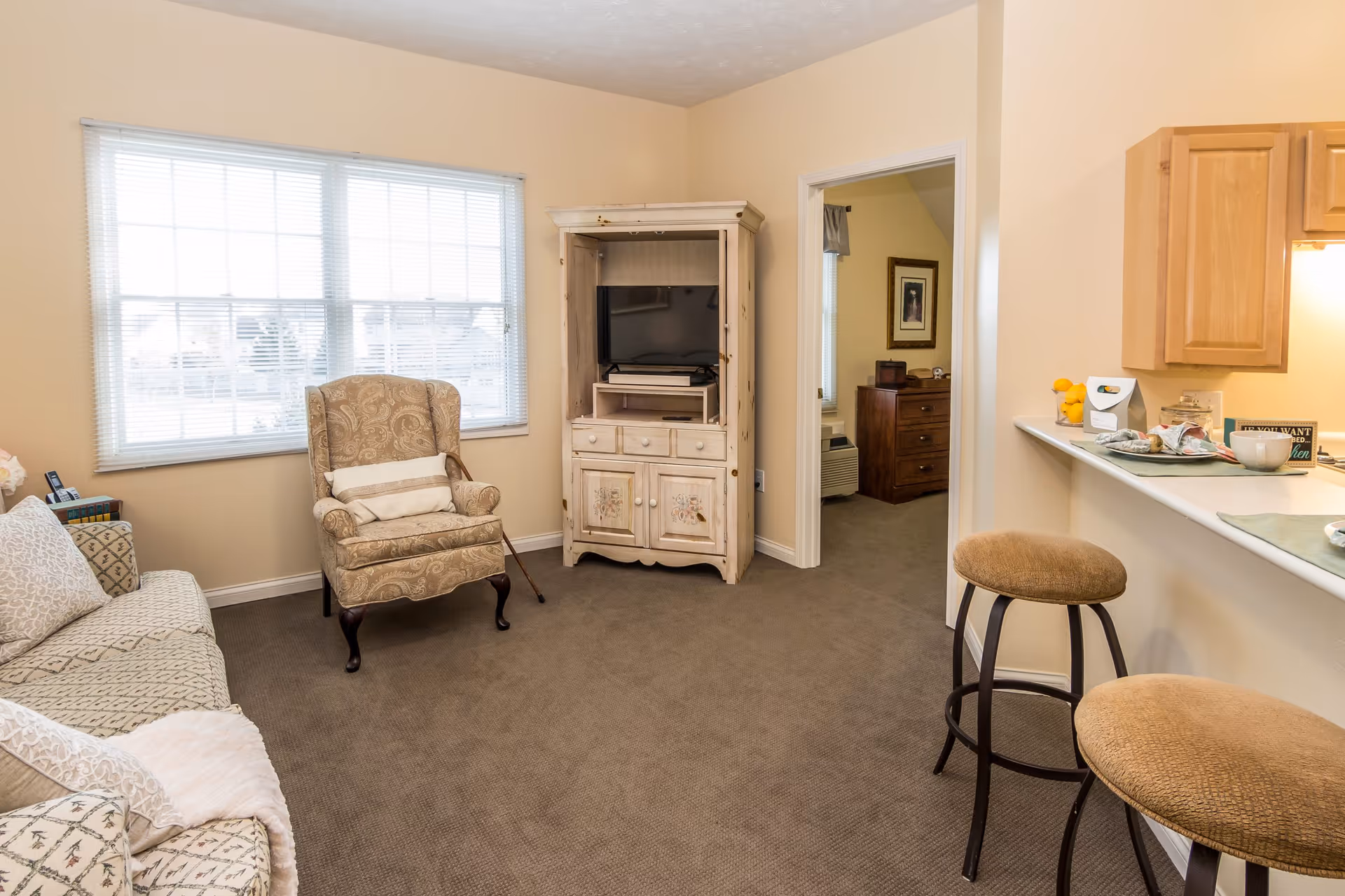 A cozy living room in an assisted living facility with a patterned armchair, a sofa with cushions, a wooden TV cabinet with a television, and a small kitchen counter with two cushioned bar stools. There is a window with blinds letting in natural light and an open doorway leading to another room with a dresser and framed artwork.