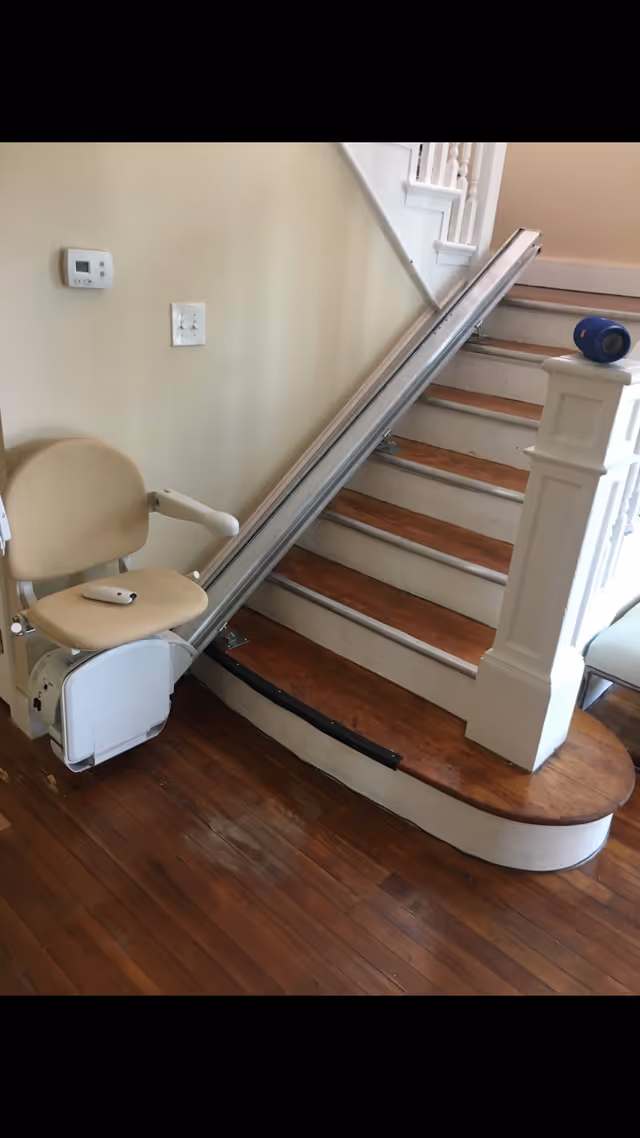 A beige stairlift installed on a wooden staircase with white risers and a white banister. The stairlift chair has armrests and a remote control placed on the seat. The surrounding area has wooden flooring and a thermostat on the wall.