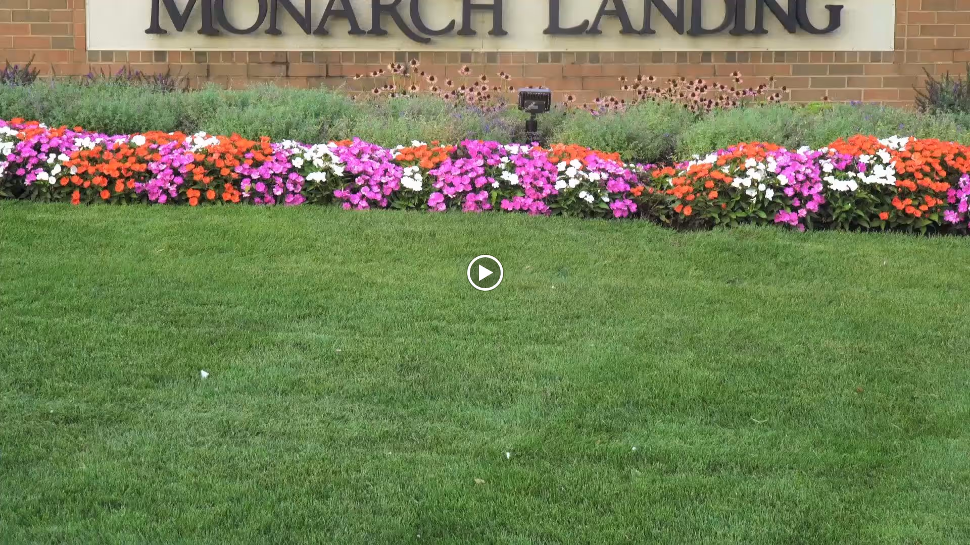 A well-maintained green lawn with a colorful flower bed of red, pink, white, and purple flowers in front of a brick wall with a sign that reads 'MONARCH LANDING'.