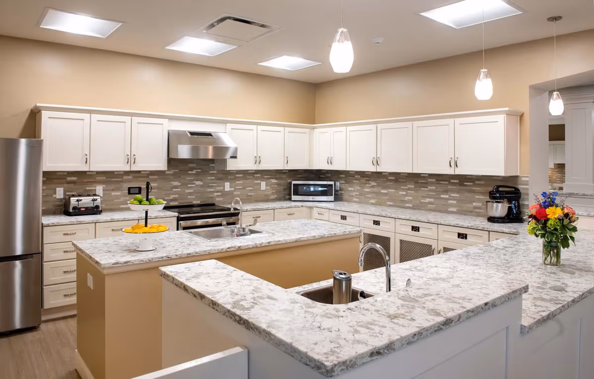 A modern kitchen with white cabinets, marble countertops, and a beige backsplash. The kitchen features a stainless steel refrigerator, stove with a range hood, microwave, toaster, and a stand mixer. There are two sinks, pendant lights hanging from the ceiling, and a vase with colorful flowers on the counter.