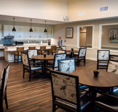 A spacious dining area with round wooden tables and chairs featuring patterned upholstery. In the background, there is a kitchen area with white cabinets, a tiled backsplash, and three pendant lights hanging over a counter with bar stools. The room has wooden flooring and neutral-colored walls decorated with framed artwork.
